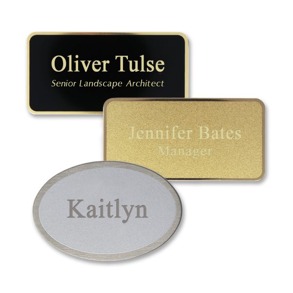 Rectangle and oval shaped black, gold, and silver metal name tags with engraved text on a matte metal face with shiny metallic underneath.